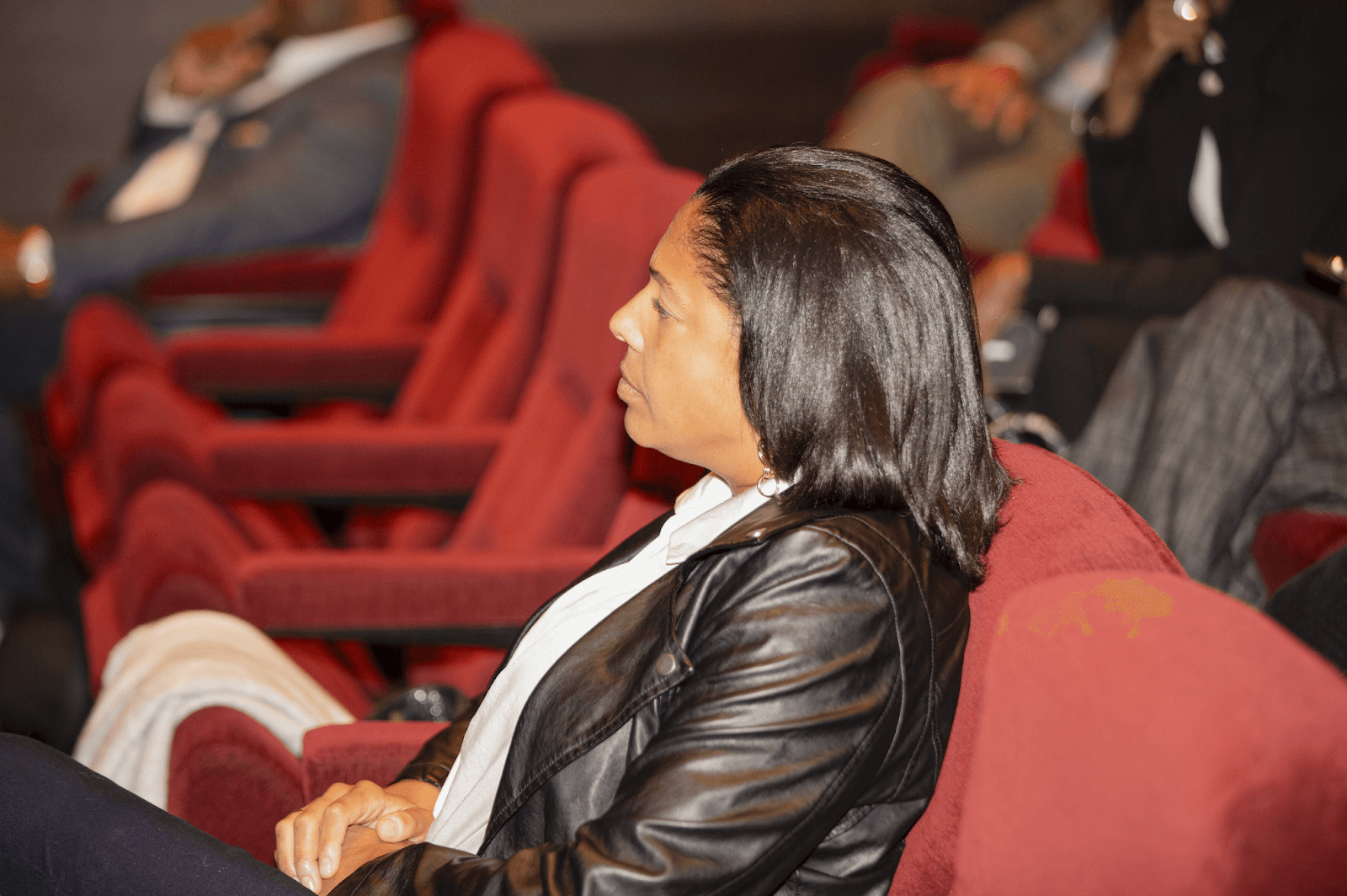 A woman wearing a leather jacket sits in a red theater chair, focused on a speaker in the foreground.