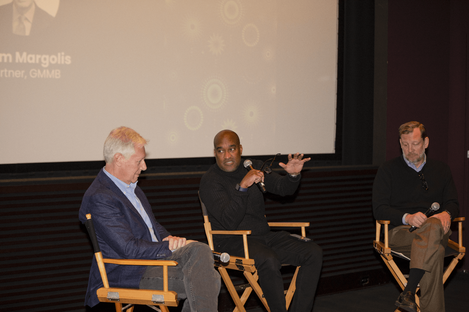 Three men on stage seated in director-style chairs, one speaking into a microphone while gesturing, participating in a panel discussion.