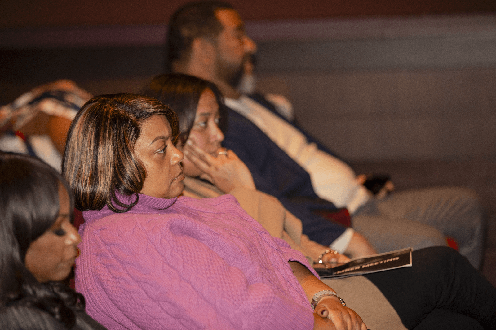 A woman in a purple sweater sits in the audience with a thoughtful expression, listening attentively to a presentation.