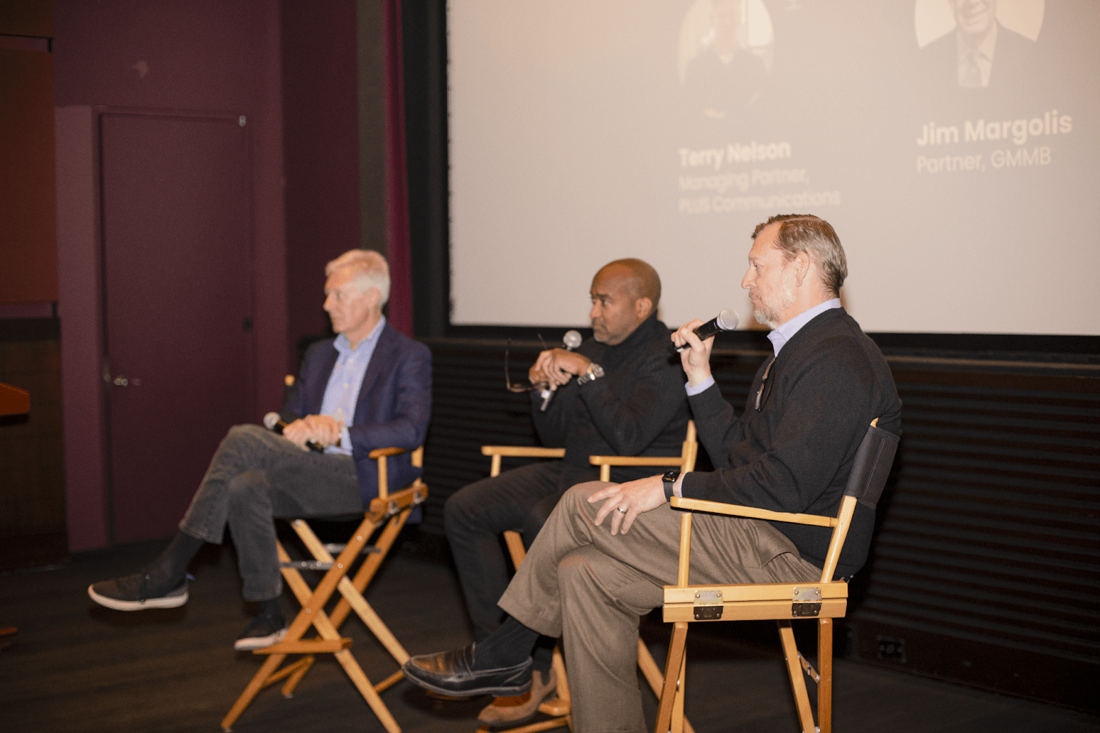 Three panelists, including one man speaking into a microphone, are seated on stage in a theater, engaging in a discussion.