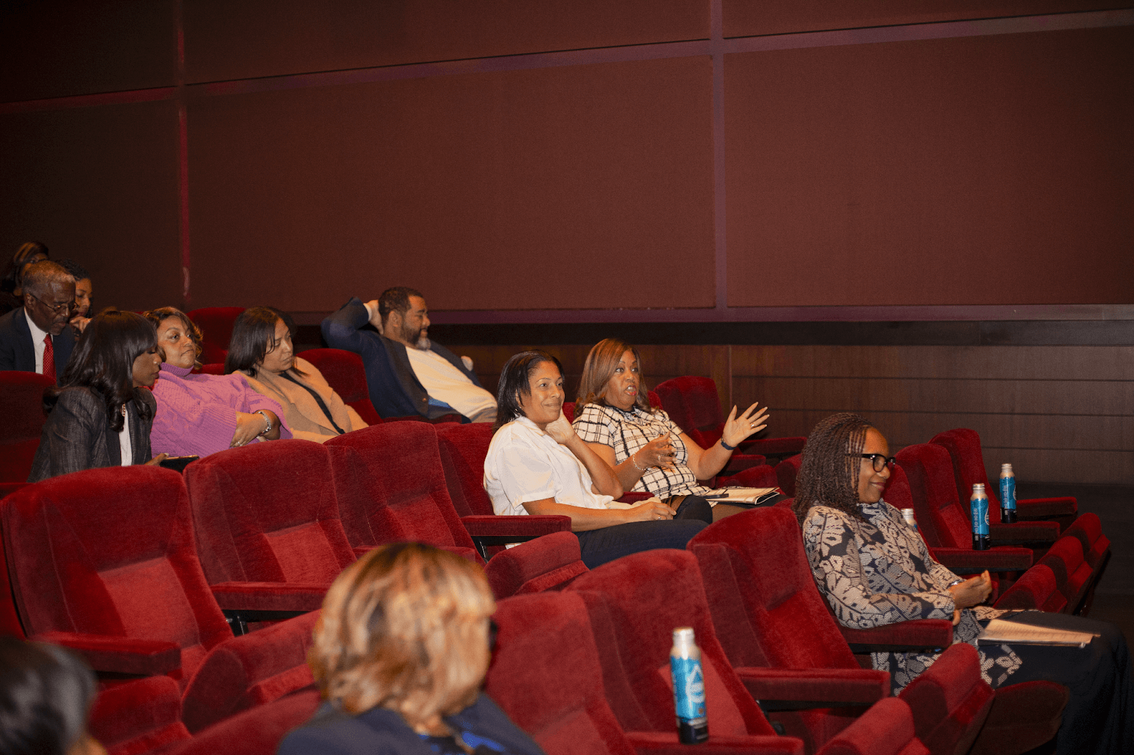 Attendees seated in red chairs listen and engage during a theater-style presentation, with one woman gesturing as she speaks.