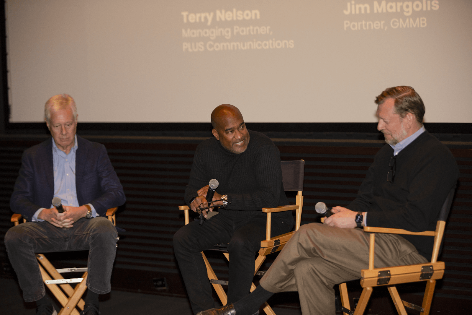 Three panelists seated on stage in director-style chairs, all holding microphones, participating in a discussion.