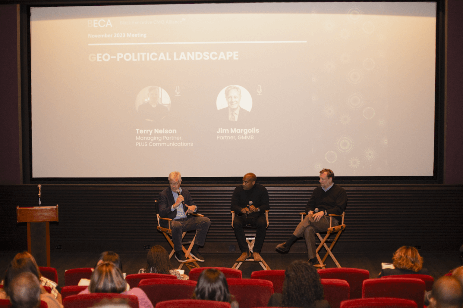 A wide shot of a theater presentation titled "Geo-Political Landscape," with three panelists seated on stage and an audience watching attentively.