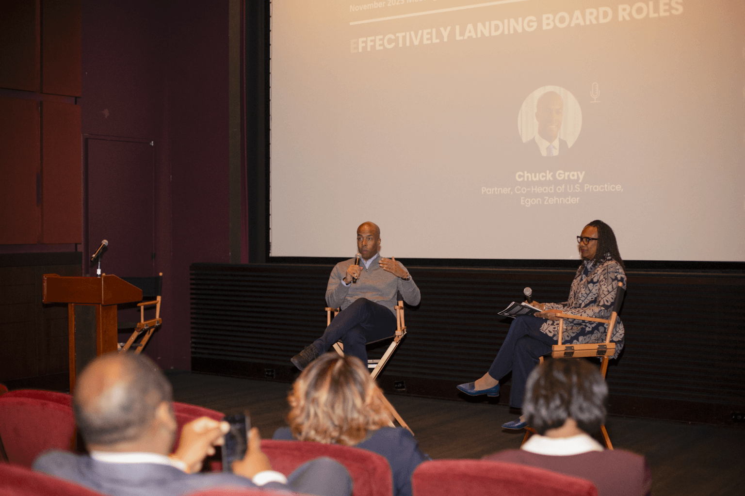 Two panelists seated on a stage, engaging in a discussion in front of a screen that displays the event's title and one speaker's name.