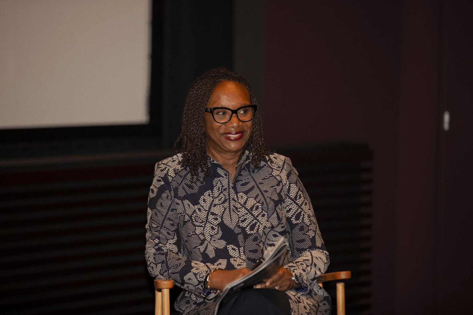 A panelist in a patterned blouse and glasses, seated and holding a microphone, smiling while participating in the event.