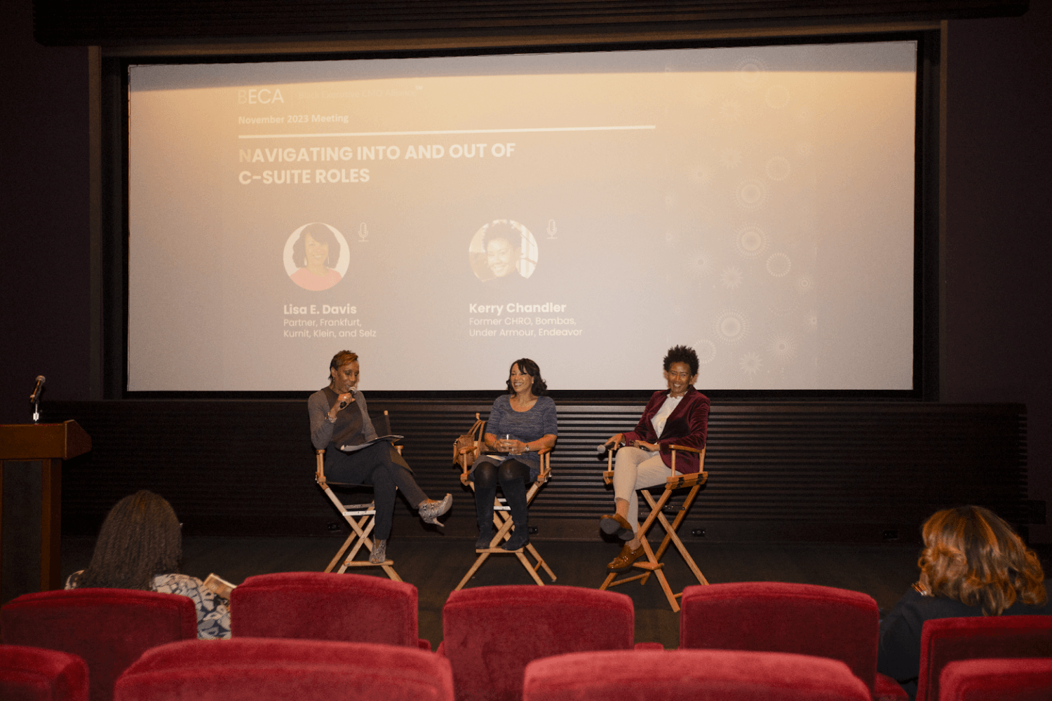 A panel discussion on stage, featuring three speakers seated in director's chairs, discussing "Navigating Into and Out of C-Suite Roles," as displayed on the screen behind them.
