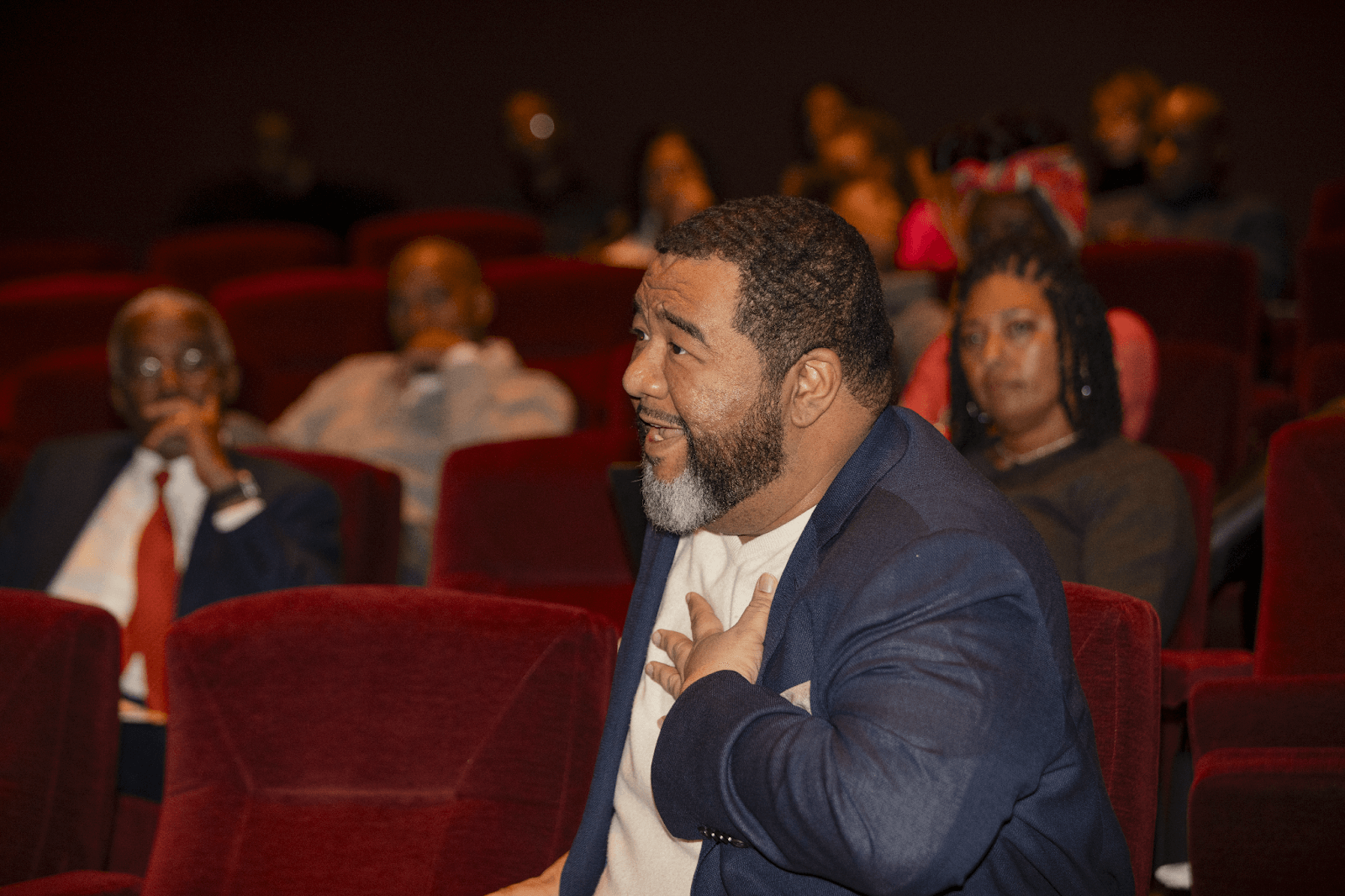 A man in a blue blazer with a white shirt animatedly speaking from the audience, with other attendees seated behind him observing.