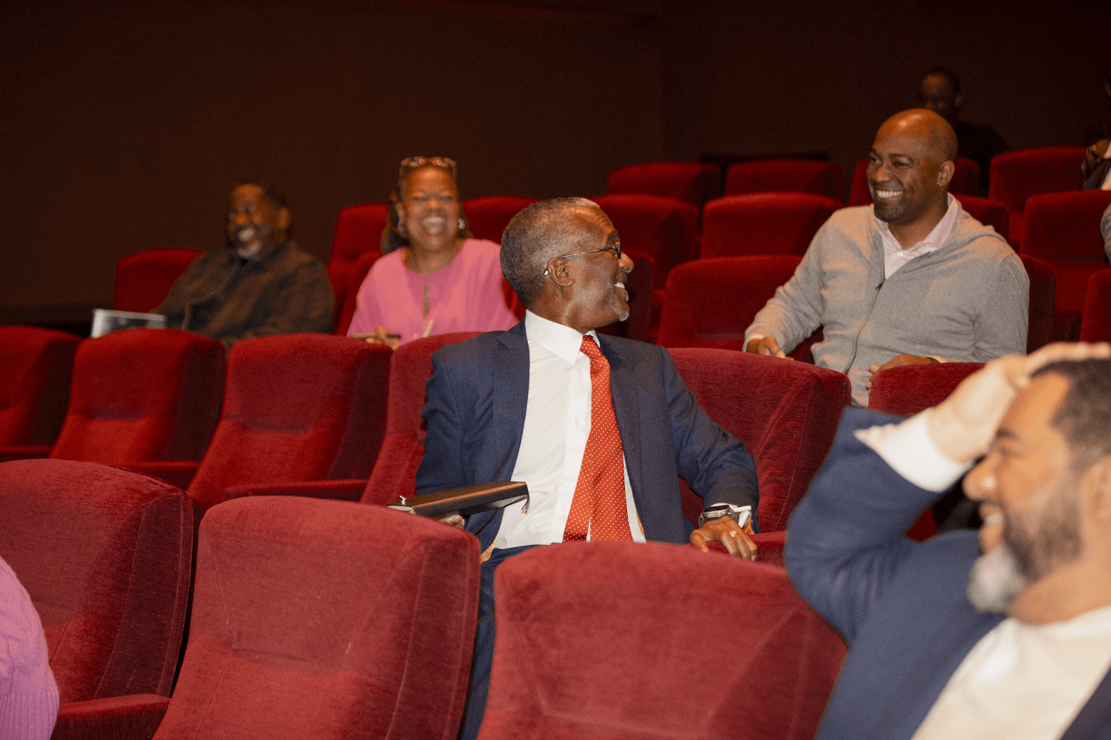 A candid moment showing a man in a suit with a red tie turning to converse with another attendee, while others in the background are smiling and chatting.
