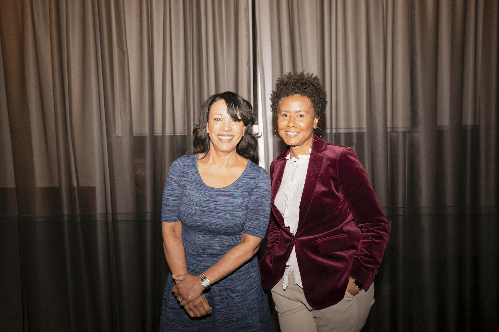 Two panelists smiling while posing together after the discussion, standing against a backdrop of neutral drapes.