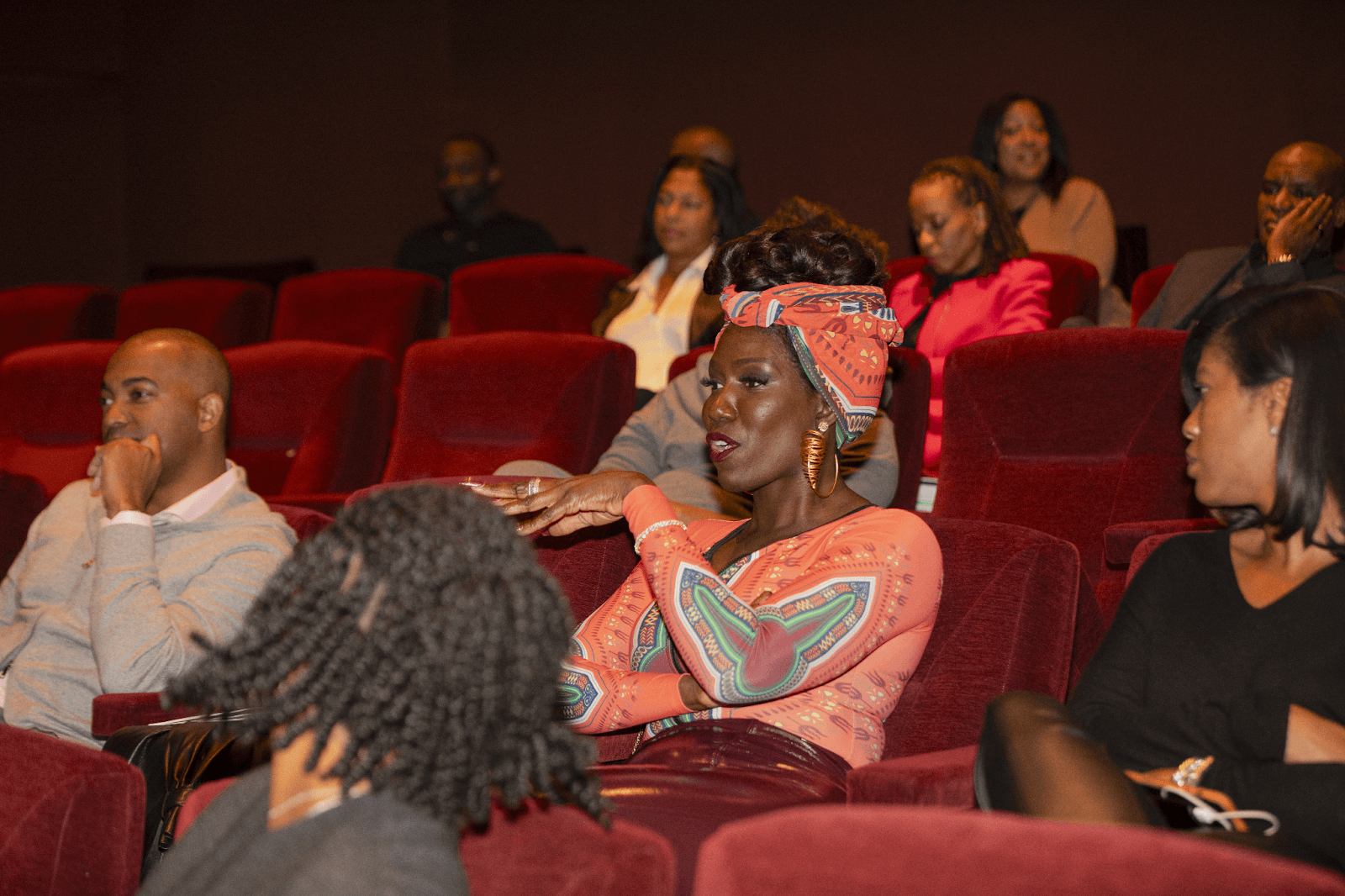 An engaged audience member in a vibrant orange outfit and matching headwrap speaks passionately during the event, surrounded by other attendees in red theater chairs.
