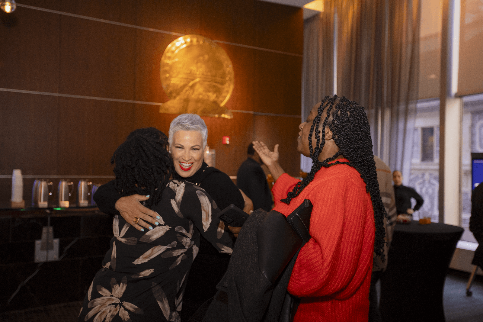 A woman with short hair warmly hugging another attendee at a social gathering.