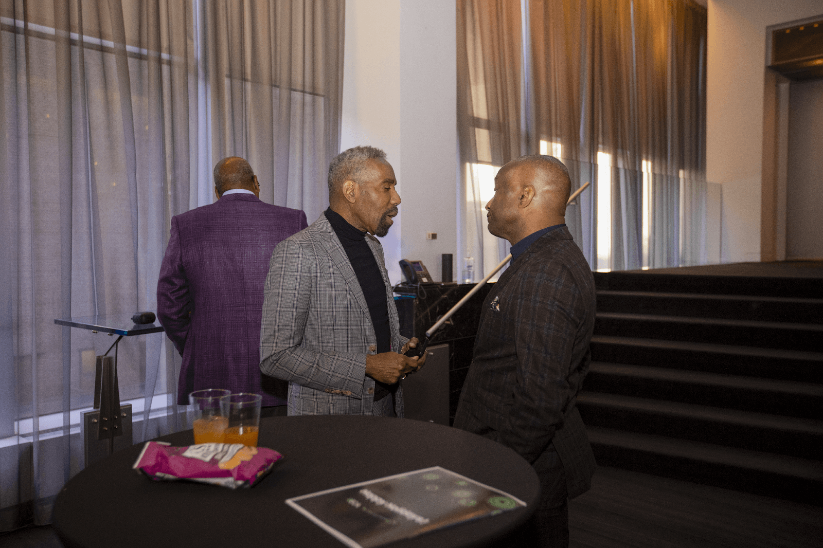 Two men in conversation at a high-top table during a professional event, with modern decor in the background.
