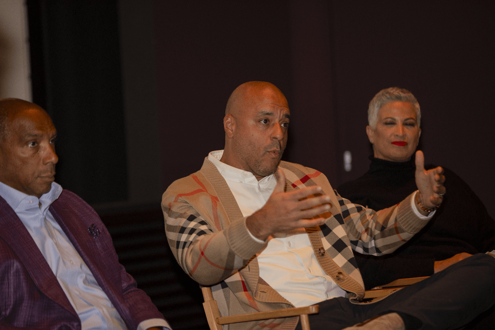 A panelist gesturing while speaking, flanked by two attentive co-panelists, in a well-lit conference setting.