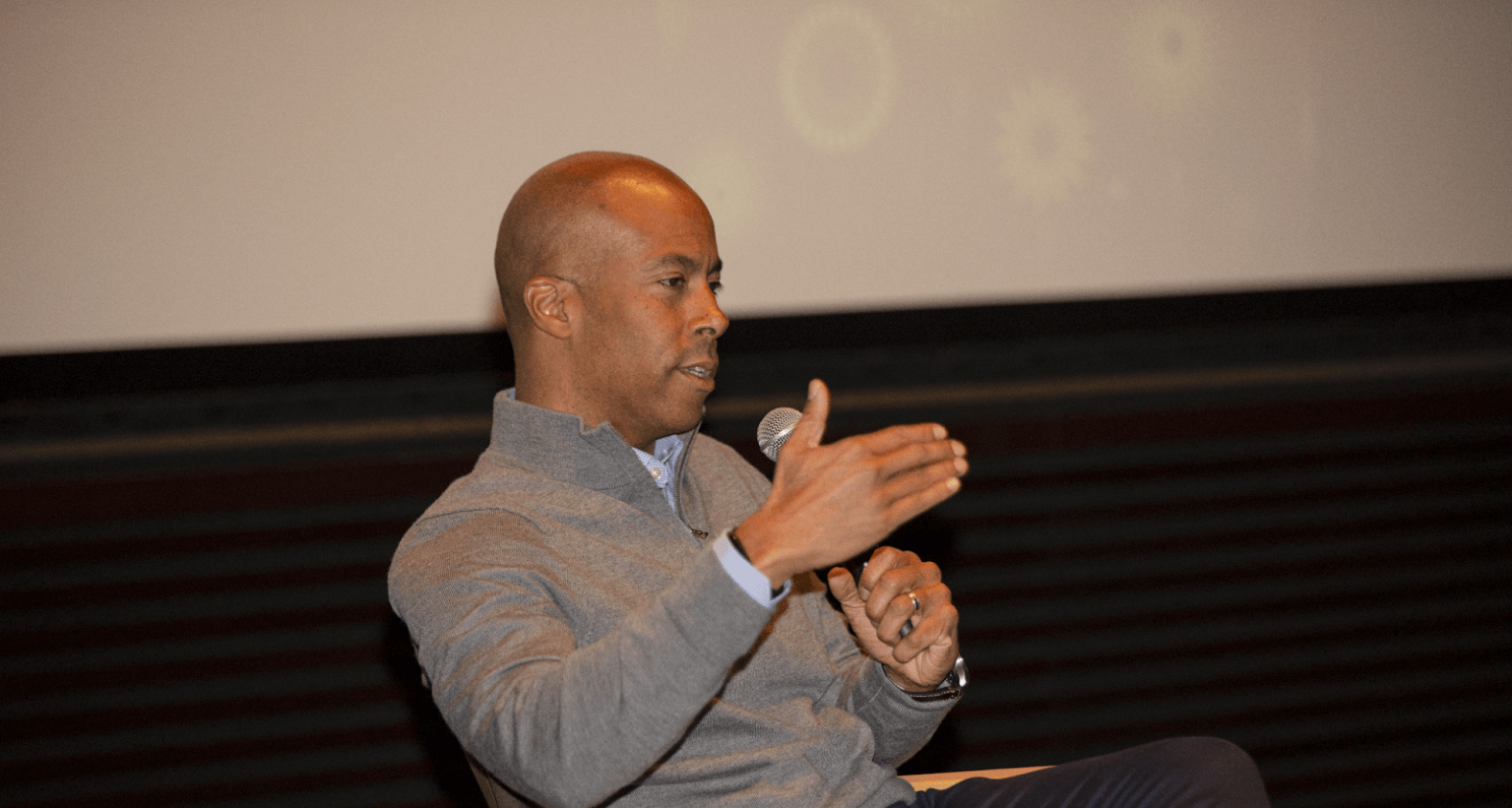 A man is seated on stage, speaking into a microphone and gesturing with his hand. He is dressed in a gray zip-up sweater over a collared shirt, appearing engaged in a discussion. The background features a large projection screen with subtle visuals, indicating a professional panel or conference setting. His focused expression suggests he is sharing insights or answering a question.