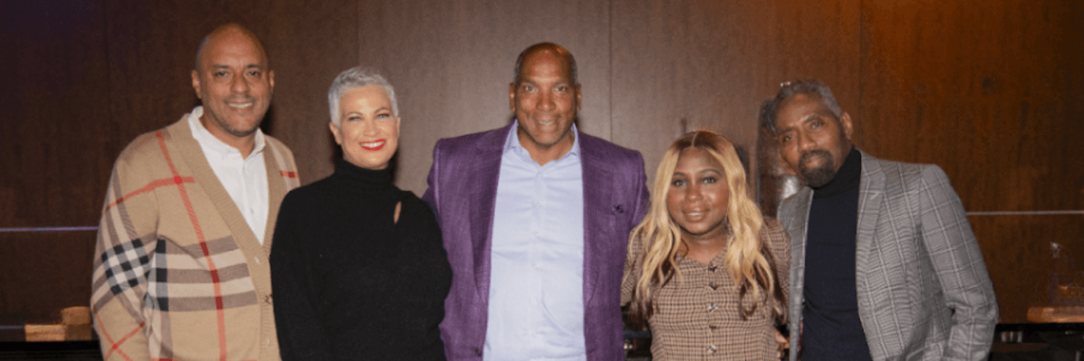 Five individuals pose together in an indoor setting, dressed in stylish and professional attire. They are smiling and standing closely, conveying a sense of camaraderie. The group includes a mix of fashion-forward outfits, including a plaid cardigan, a sleek black dress, a purple blazer, and patterned jackets. The warm wooden backdrop suggests a sophisticated networking or leadership event.