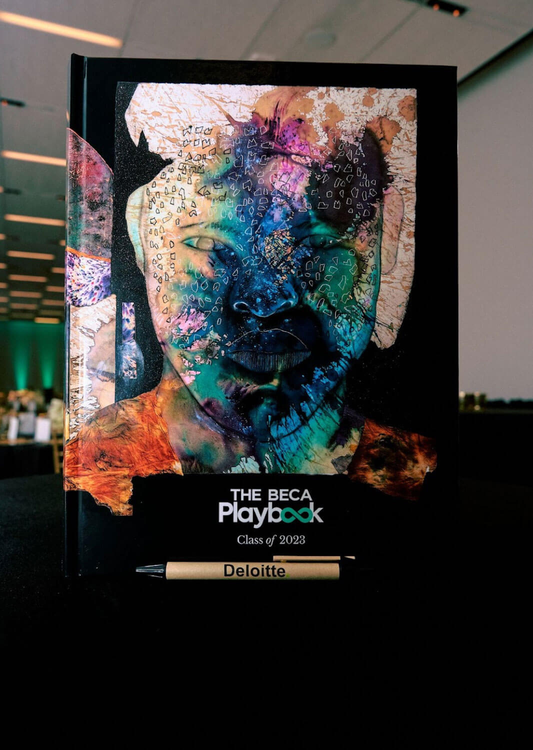 A colorful artistic cover of "The BECA Playbook: Class of 2023," positioned upright on a table with a Deloitte-branded pen in front.
