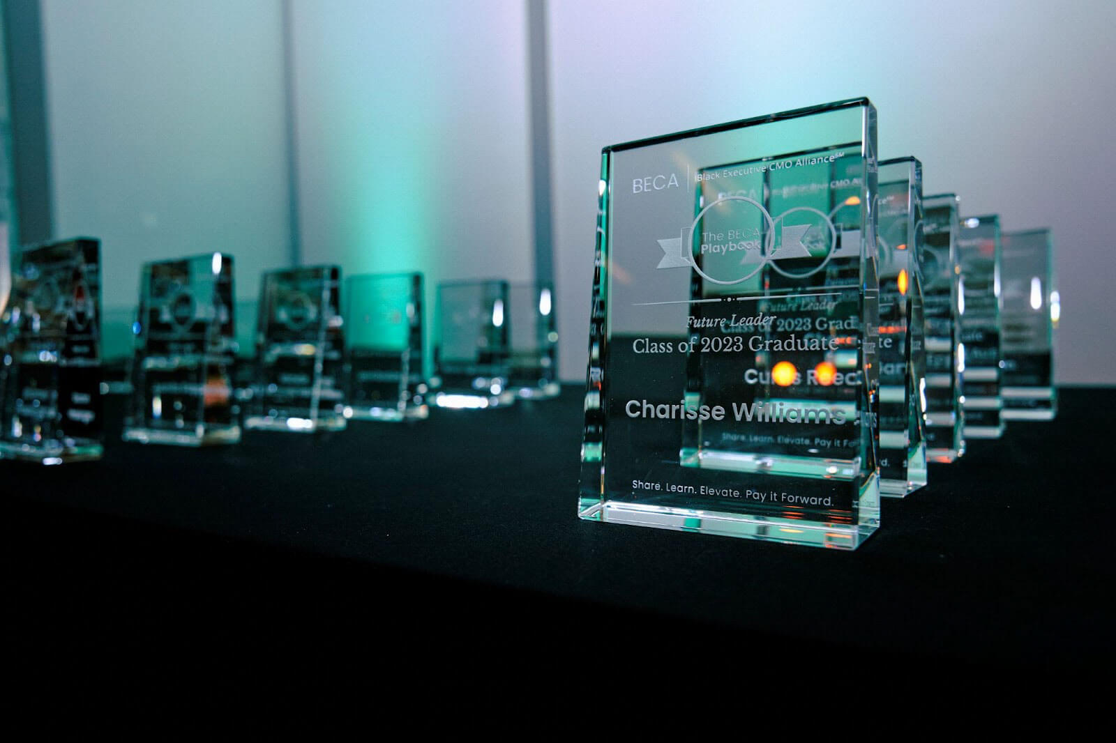 A row of glass awards displayed on a black table, each engraved with "BECA Black Executive CMO Alliance," "The BECA Playbook," and personalized details, including the name "Charisse Williams" and the title "Future Leader 2023 Graduate."
