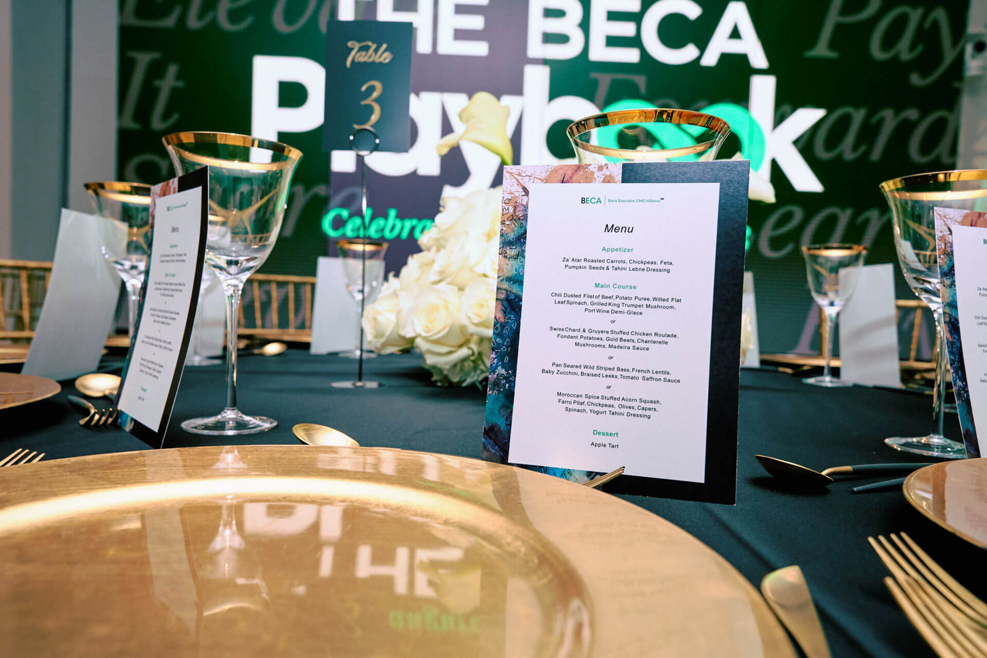 A table setting with elegant gold-rimmed glasses and a detailed menu for the BECA Playbook event, featuring an appetizer, main courses, and dessert options.