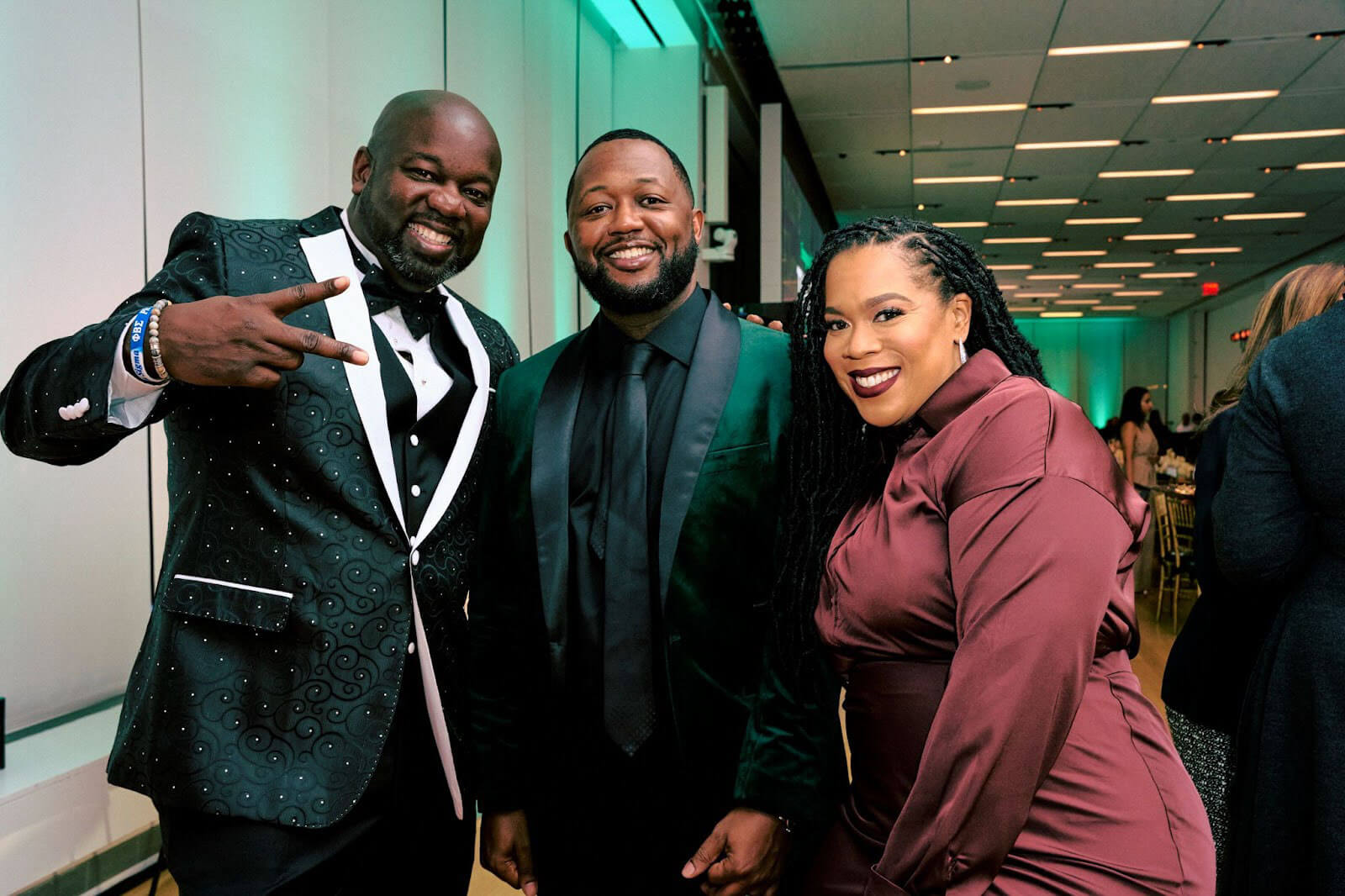 Three attendees dressed in formalwear posing for a photo, two men in tuxedos and a woman in a burgundy dress.