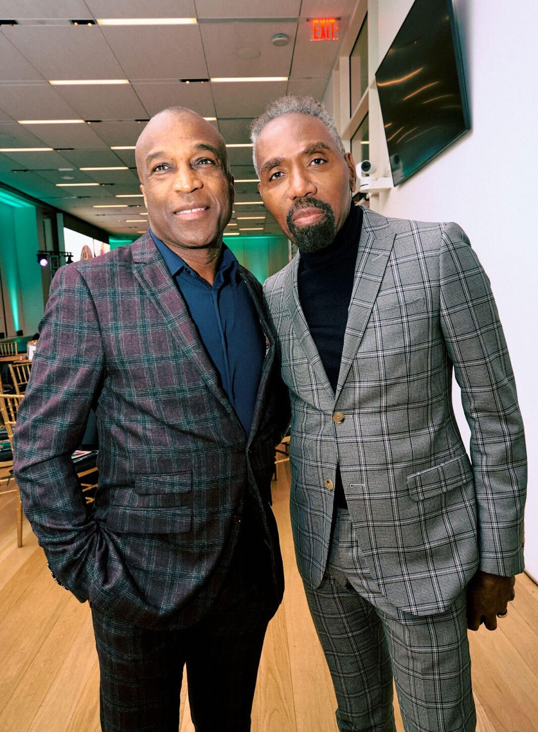Two men in plaid suits posing together in a formal setting with bright lighting and tables in the background.