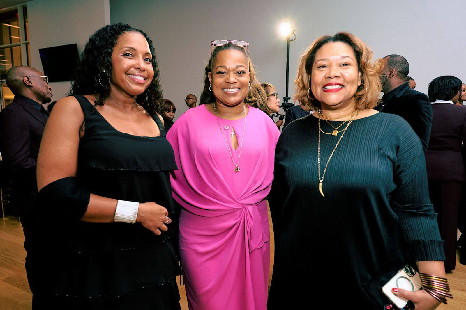 Three women posing together, one in a black dress, another in a bright pink outfit, and the third in a black dress with gold accessories, against a lively indoor backdrop.