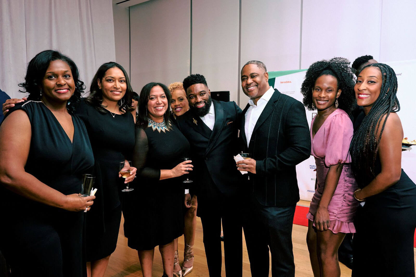 A group of eight people dressed in formal wear, smiling and holding drinks in a well-lit event space with a white curtain background.
