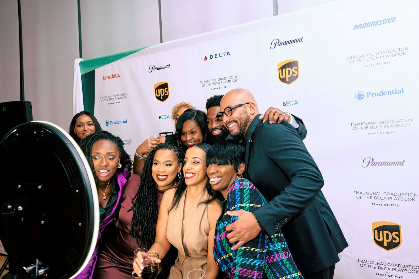 A group of people huddled together, smiling for a group photo in front of a white backdrop featuring corporate logos and event branding.