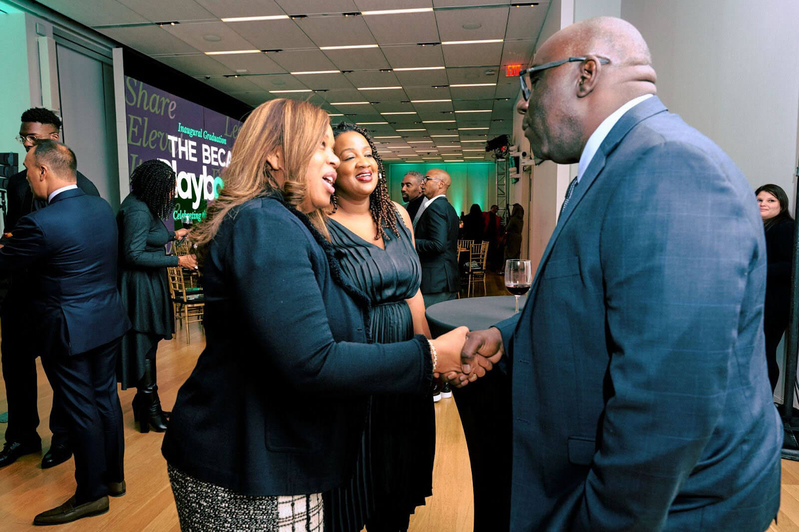 A man and a woman shaking hands at a formal event, with others engaging in conversation and event banners in the background.