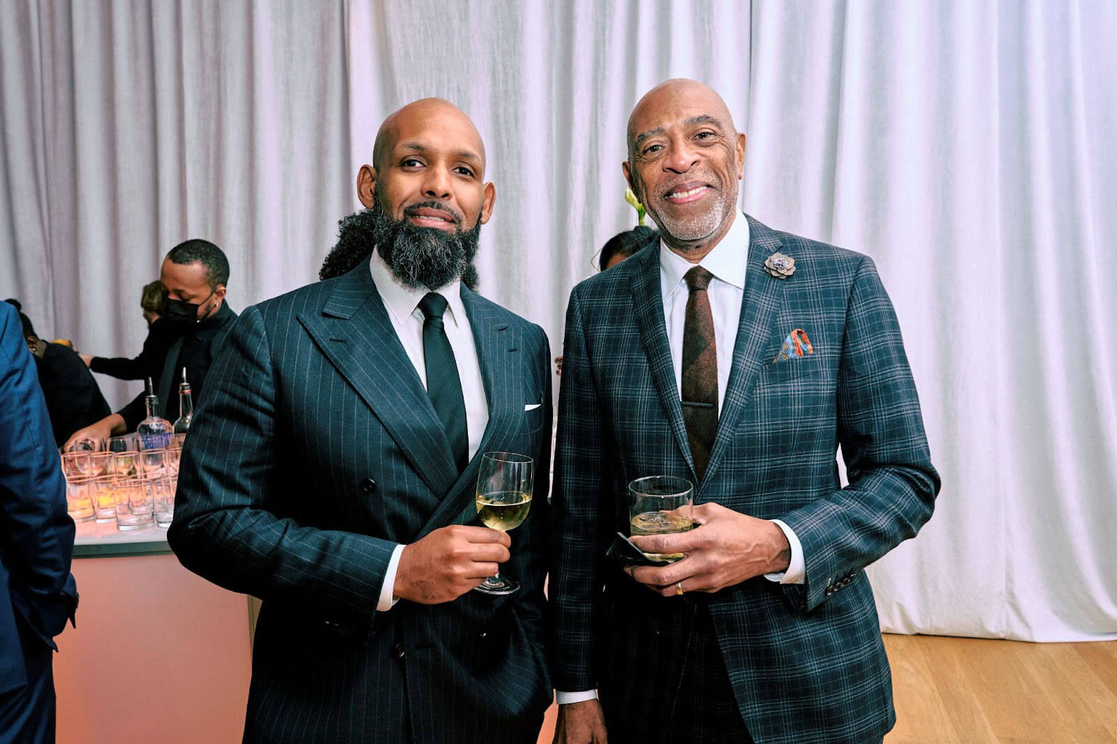 Two men dressed in formal suits, one with a neatly groomed beard and the other with a flower lapel pin, holding drinks and smiling in front of a white curtain backdrop at an elegant event.