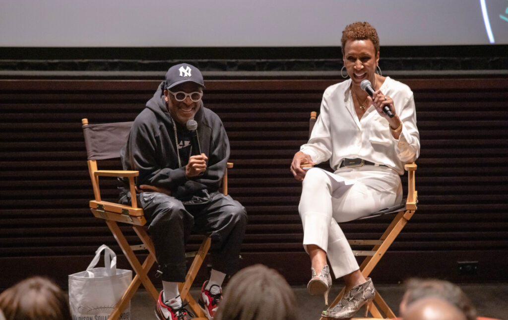 Two people seated on directorâs chairs hold microphones on a stage. The man, wearing a dark hoodie, sunglasses, and a baseball cap, smiles while speaking. The woman, dressed in a white blouse and pants, also smiles as she addresses the audience.
