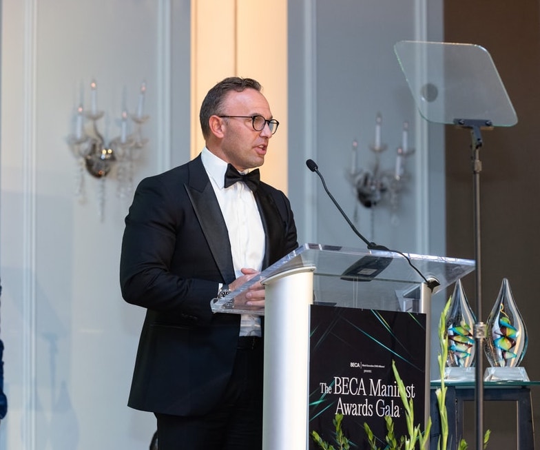 A man in a black tuxedo and glasses speaks from a clear podium labeled “The BECA Manifest Awards Gala,” with glass award trophies visible beside him.
