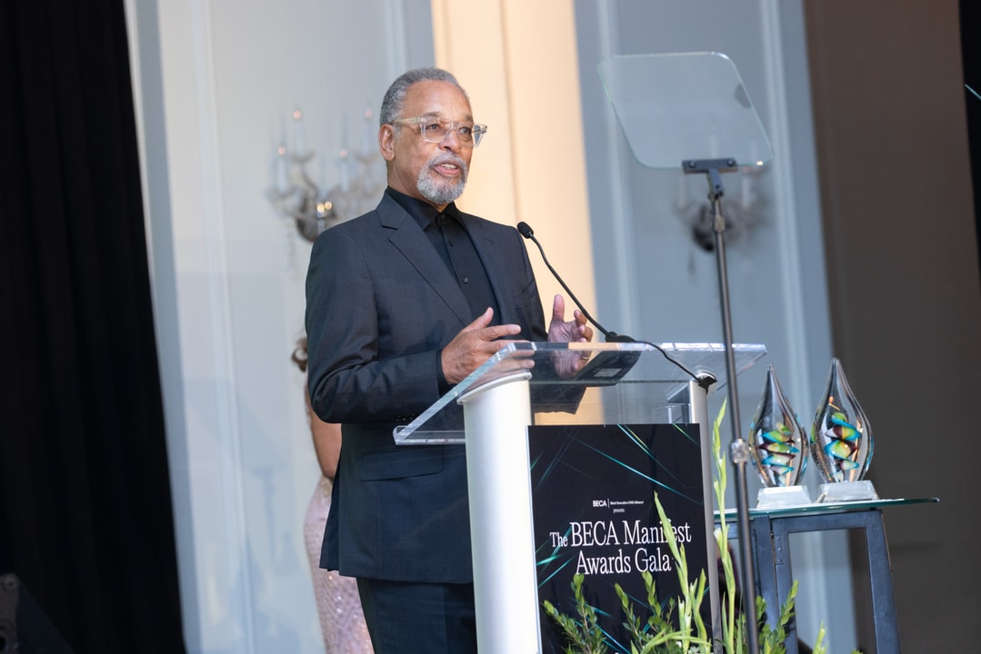 A man in a dark suit and glasses speaks at a clear podium labeled “The BECA Manifest Awards Gala,” with glass award trophies displayed to his right.