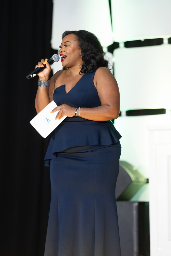 A woman in a navy, peplum-style gown holds a microphone and an event program, smiling as she speaks onstage before a black backdrop.