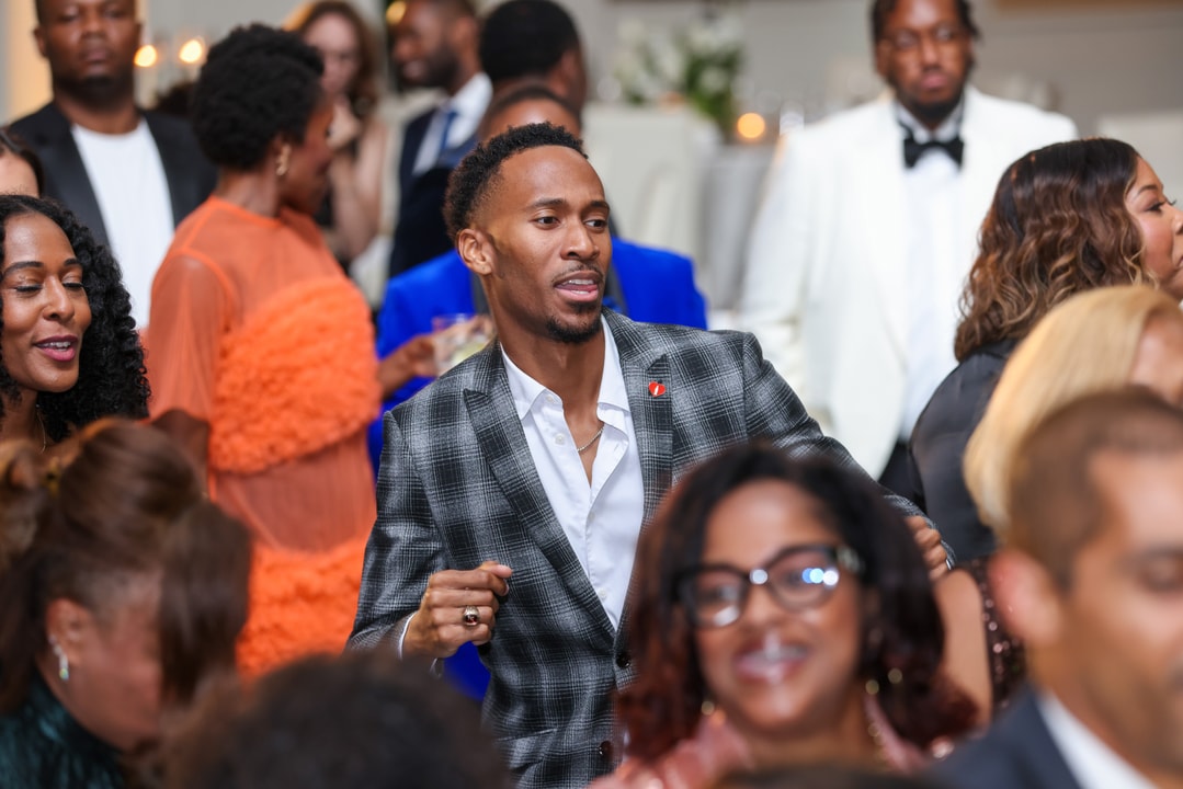 A man in a stylish gray plaid suit dances with a lively expression at a crowded event, surrounded by elegantly dressed guests enjoying the celebration.