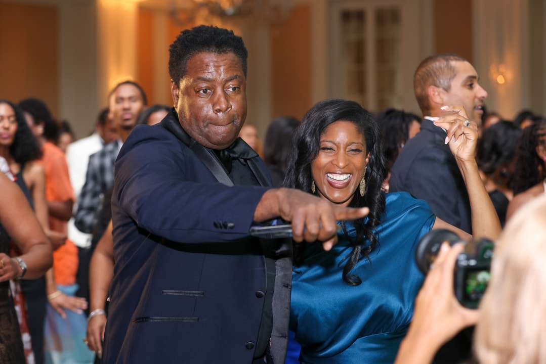 A man in a navy blue suit energetically points towards the camera while dancing, alongside a woman in a blue satin dress who is laughing joyfully at a lively event surrounded by other guests.
