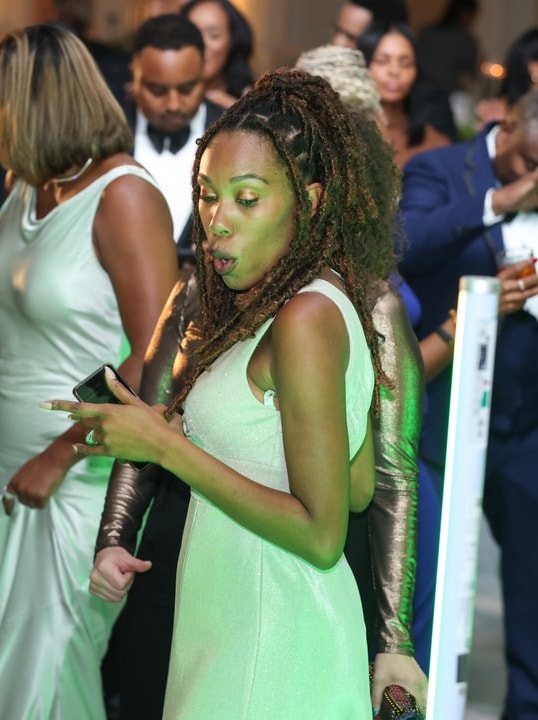 A woman in a light-colored dress dances with an excited expression, surrounded by elegantly dressed guests at a formal event, under green lighting.