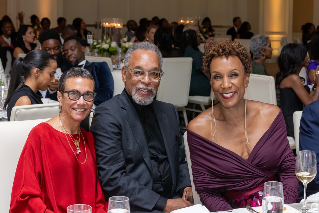 Three elegantly dressed individuals pose for a photo at a gala, smiling warmly. The woman on the left wears a red dress with gold accessories, the man in the center wears a black suit with glasses, and the woman on the right wears a burgundy off-the-shoulder gown with long earrings. The background shows other guests, candles, and floral centerpieces, creating a lively and sophisticated atmosphere.