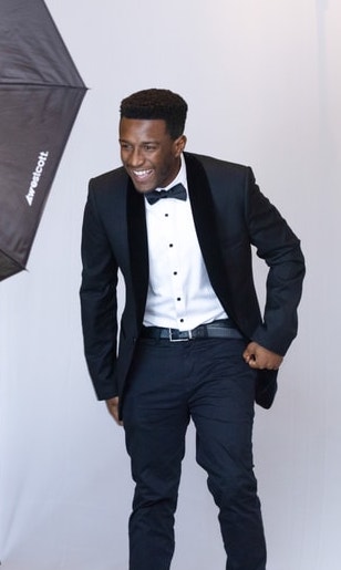 A young man in a black suit, white shirt, and bow tie leans forward with a broad smile, posing against a white studio backdrop with a photography light visible on the side.