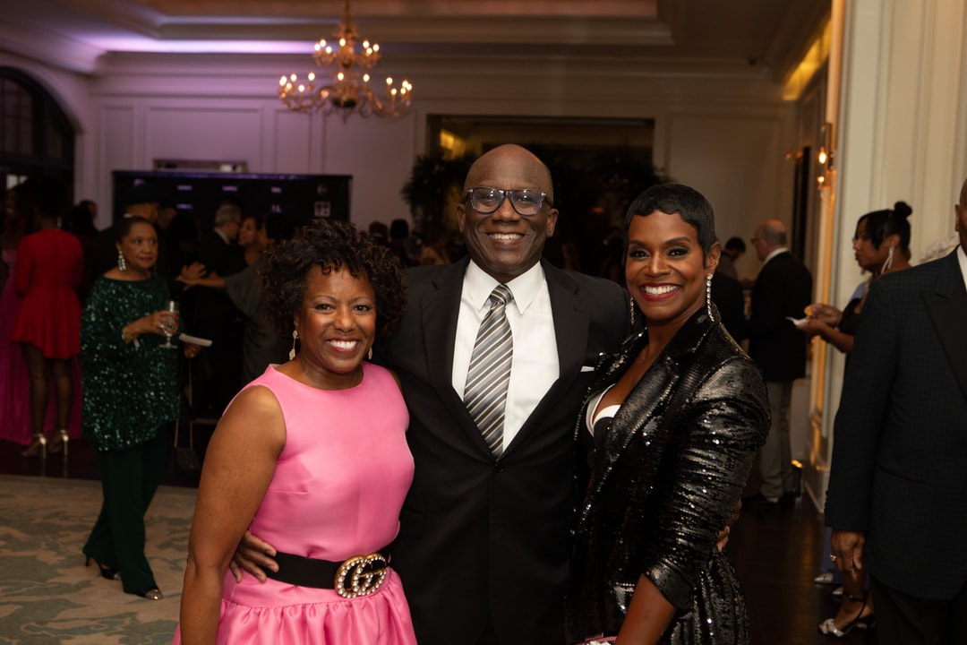 A woman in a bright pink dress, a man in a suit and a woman ina black glittery dress smiling and posing for a photo.