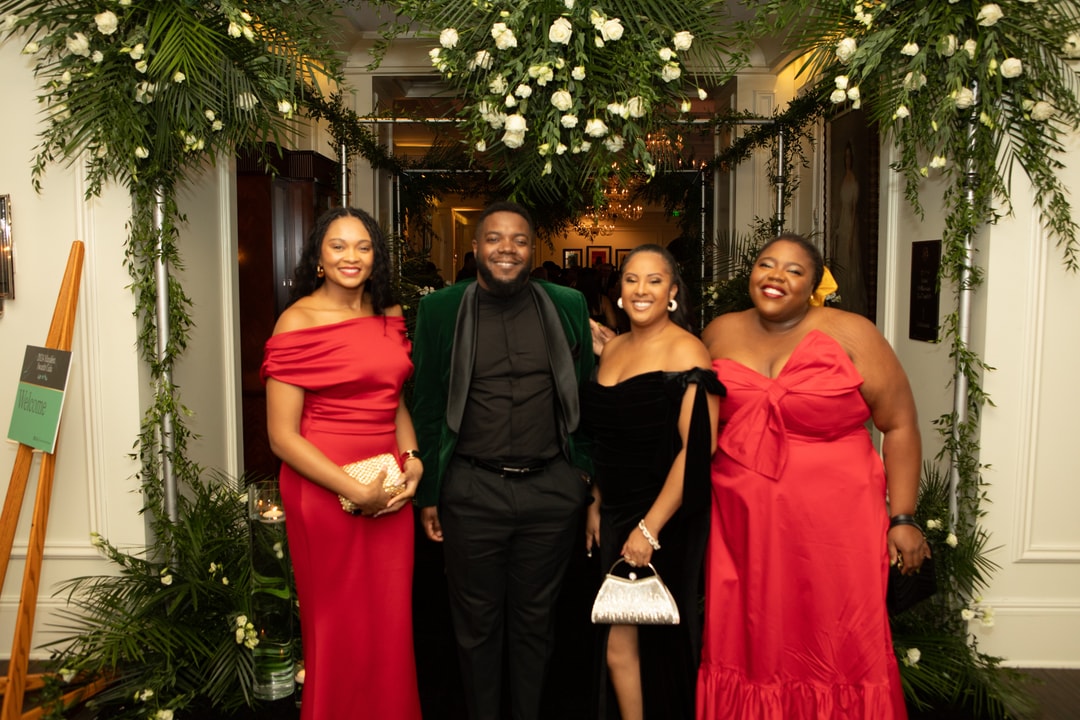 A woman in a red dress, a man in a black suit, a woman in a long black dress, and another woman in a red dress posing together at a gala event. They are standing in front of a floral backdrop in a grand hall.
