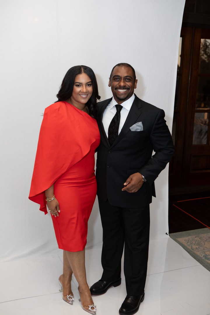 A woman in a bright red, cape-style dress stands beside a man in a black suit and tie; both smile at the camera against a white backdrop.