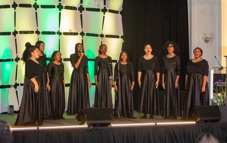 A group of singers in matching black gowns perform onstage, one holding a microphone, against a backdrop lit in green at a formal event.