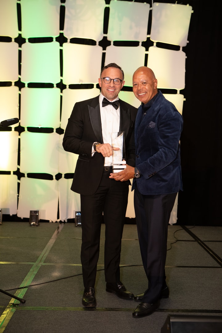 Two men in formal attire stand on a stage with a lit geometric backdrop. The man on the left, wearing a tuxedo and bow tie, holds a glass award, while the man in the navy blazer congratulates him.