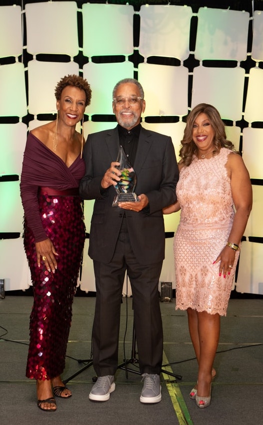 Three people stand on a stage with a lit geometric backdrop. The man in the center, dressed in black and holding a glass award, is flanked by two women wearing elegant gowns, all smiling at the camera.