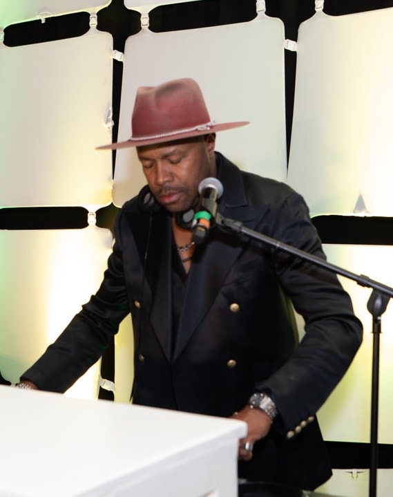 A musician wearing a stylish hat and black double-breasted jacket performs at a keyboard on stage, with a microphone positioned in front of him against a geometric-lit backdrop.