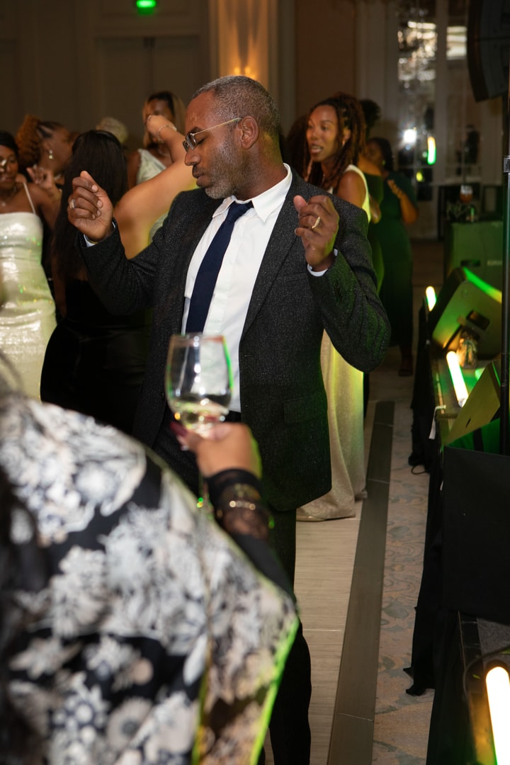 A man in a dark suit and glasses dances with enthusiasm at a formal event, surrounded by elegantly dressed guests, with green lighting in the background.