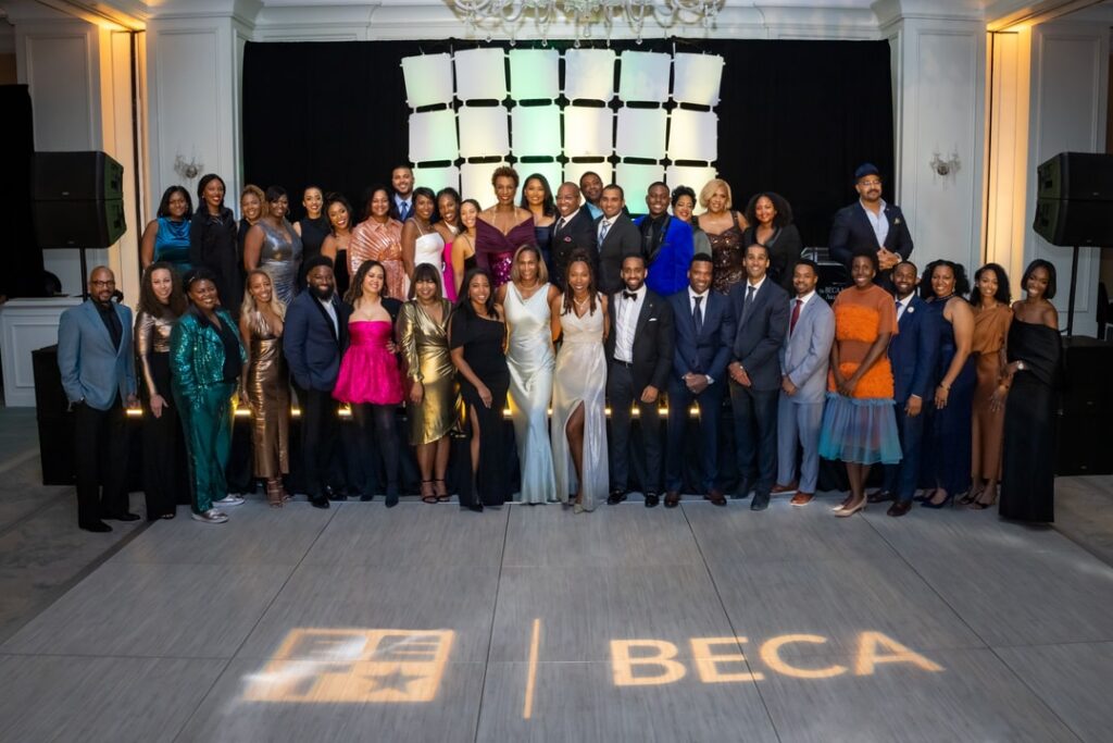 A large group of elegantly dressed people poses on a ballroom stage at the BECA 2024 Gala. The floor displays the BECA logo, and the setting features a stylish backdrop with warm lighting.