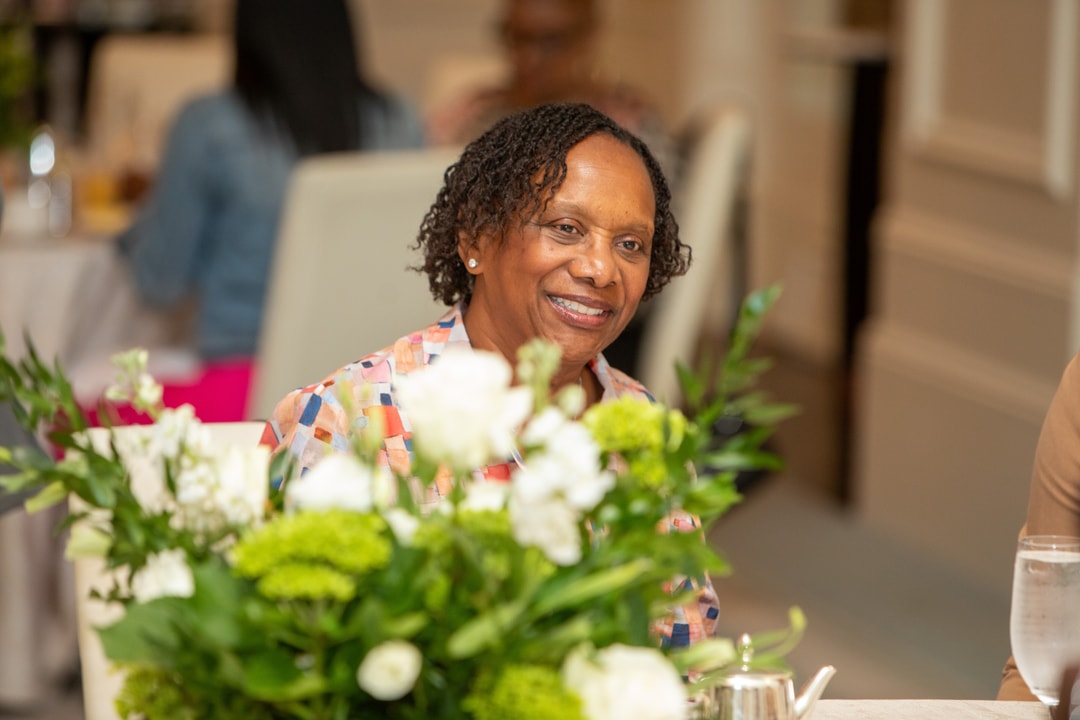 A woman in a colorful patterned blouse smiles while seated at a table with a floral centerpiece, appearing engaged in the event.