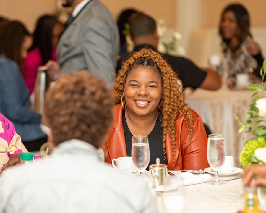 A young woman with curly hair, wearing a black outfit and an orange blazer, smiles brightly while seated at a round table, enjoying the conversation.