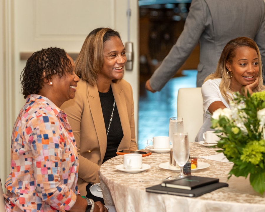 Three women sit at a round table, engaged in conversation and smiling. The table is set with coffee cups, notebooks, and a floral centerpiece, and a man in a suit is seen walking in the background.
